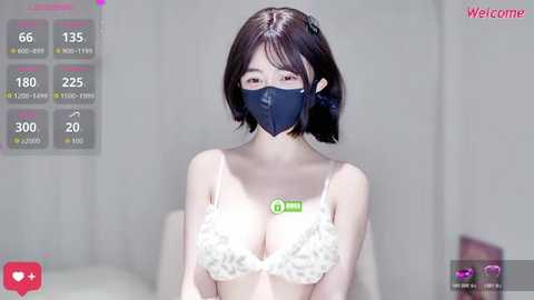 A video of an Asian woman with short black hair, wearing a white lace bra and a black face mask, standing against a blurred, light-colored background.