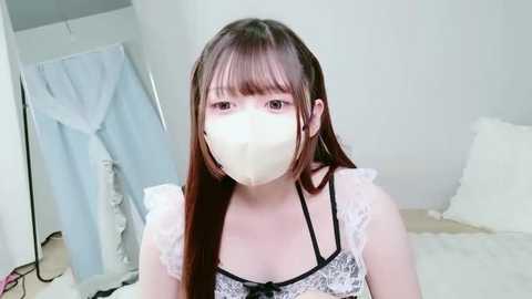 Video of an East Asian woman with long, straight brown hair, wearing a white lace-trimmed bralette, face mask, and sitting on a bed in a plain, white room.