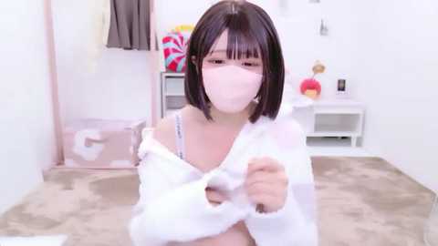 Media: Video of a young Asian woman with short black hair and a white face mask, wearing a white, fluffy jacket, sitting in a clean, white room with a pink stuffed toy.