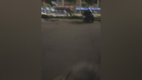 A blurry nighttime photo of a dark-skinned person on a bicycle in an urban street with dimly lit storefronts and a blurred figure walking behind.