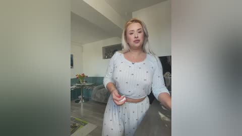 Video of a plus-sized woman with blonde hair in light blue pajamas, standing in a modern living room with white walls, gray couch, and floral arrangement on a table.