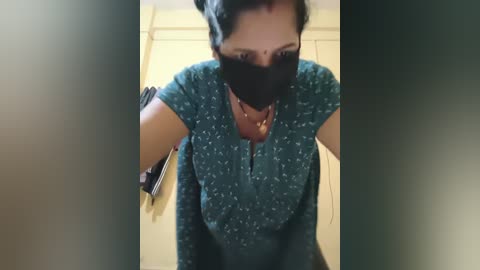 A video shows a South Asian woman with a medium complexion, short dark hair, wearing a black mask, teal-patterned shirt, and a yellow sari, leaning forward, possibly in a bathroom.