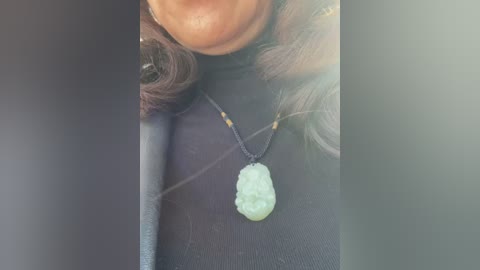 Media: Video of a person wearing a black turtleneck sweater and a light green pendant necklace with a textured stone. The background is blurred, with a dark edge on the left and a lighter edge on the right.