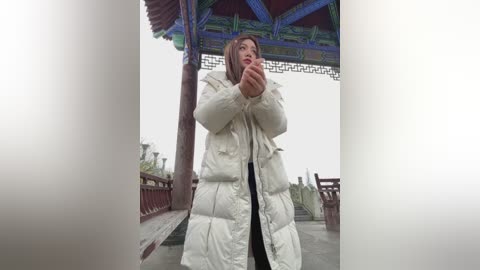 Video of a young East Asian woman in a white puffy coat, standing in a traditional wooden pavilion with red and blue accents, holding a cup of tea.