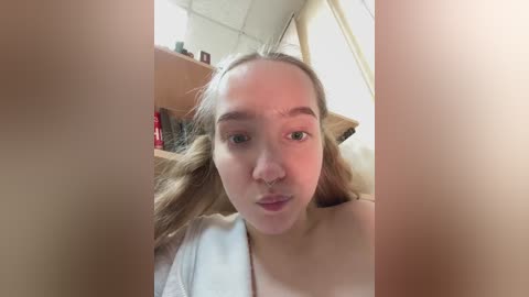 Video of a young, light-skinned woman with long blonde hair, wearing a white robe, sitting indoors with a neutral expression, surrounded by a blurry background with shelves and objects.