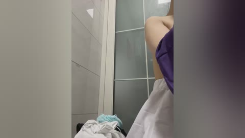 A video of a person in a white shirt and purple top, standing in a narrow, tiled bathroom with a glass shower door and a pile of clothes on the floor.
