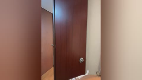 Media: Video of a narrow, dimly lit hallway with wooden doors on either side, one with a silver doorknob. The walls are beige, and the floor is light brown.