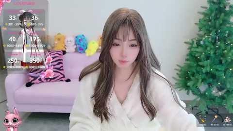 Video of an Asian woman with long brown hair, wearing a white robe, sitting in a room with a pink sofa, zebra-patterned pillow, and a green Christmas tree.