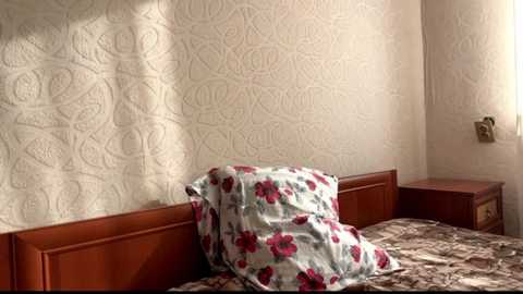 Video of a modest, vintage bedroom with a wooden bed frame and floral bedspread, adorned with a white pillow and red rose-patterned pillowcase. The wallpaper features a light beige, ornate floral design. The room is softly lit, giving it a cozy, nostalgic atmosphere.