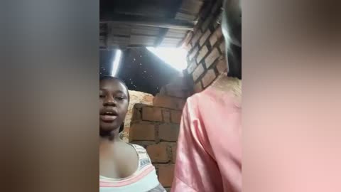 Video of a young Black woman with braids, wearing a pink satin robe, standing in a dimly lit room with exposed brick walls and a fluorescent light.