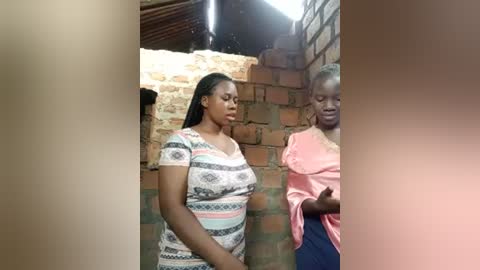 Video of two African women in a rustic, brick-walled room. One woman with braided hair wears a patterned dress, while the other in a pink blouse and blue skirt stands by her side.