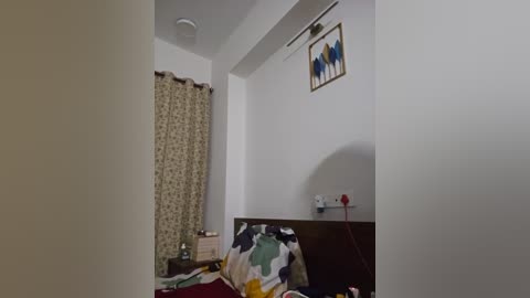Video of a small, sparsely furnished bedroom with white walls, a single bed with floral bedding, a curtain with a floral pattern, and a painting of three people on the wall.