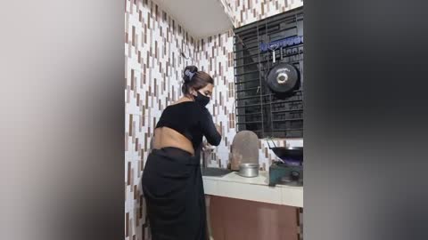 A video depicts a woman with dark hair in a bun, wearing a black mask and top, washing dishes in a small, tiled kitchen with a barred window and white and brown tiled walls.