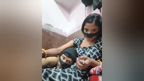 Video of a woman with a child in a small, dimly lit bathroom; the woman wears a black face mask and a black headscarf, while the child has a yellow wristband.