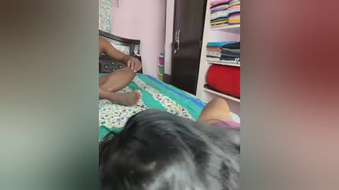 Media: Video of a person in a hospital bed with a black dog sitting beside them, viewed from the dog's perspective. The room has pastel walls, a dark wooden dresser, and a shelf with neatly folded clothes.