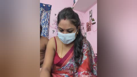 Video of an Indian woman in a red sari, wearing a blue mask, listening to earphones. Background features pastel pink walls with framed pictures, and a blue curtain.