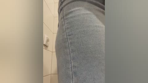 Video of a person's lower body in light gray jeans, viewed from the side. The background features beige tiled walls with a white light switch visible. The image focuses on the tight fit of the jeans.