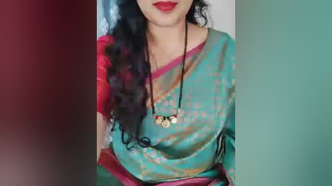 Video of a woman with long, curly black hair, wearing a teal saree with pink borders and a black necklace.