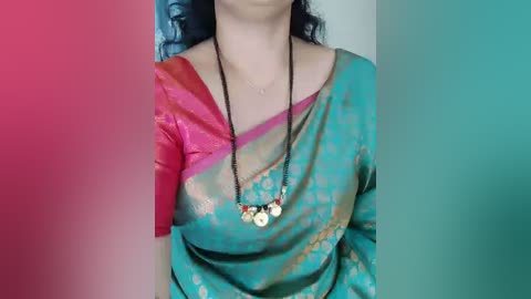 A video of a woman wearing a vibrant, teal sari with a pink and gold border, adorned with a multi-colored beaded necklace. The background features a gradient of red and teal.