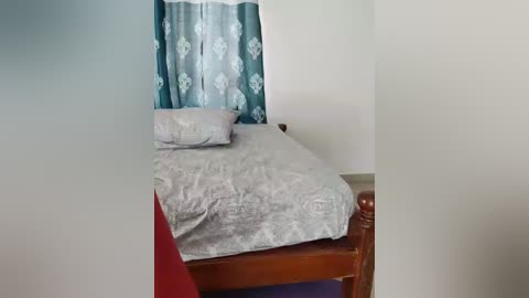 A video of a simple bedroom with a single bed covered in light grey bed sheets, adorned with a patterned pillow, and turquoise curtains with floral designs. The room has white walls and a wooden bed frame.