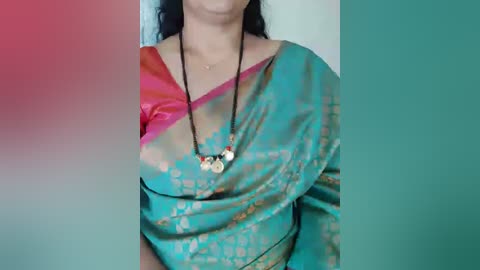 A video of a woman wearing a colorful saree with pink, green, and teal patterns, black beaded necklace, and black hair. The background is blurred in teal and red hues.