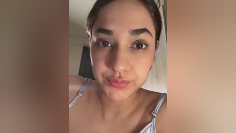 Video of a young woman with light brown skin, dark brown eyes, and dark hair pulled back. She wears a blue and white striped tank top. The background is blurred, indoor, neutral-toned.