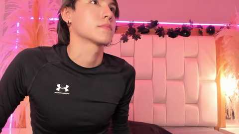 Video of a young woman with short black hair, light skin, wearing a black Under Armour shirt, in a modern, pink-lit bedroom with a white headboard and floral decor.
