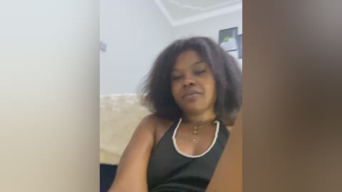 A video of a Black woman with shoulder-length, curly black hair, wearing a black sleeveless dress and pearl necklace, sitting on a cream-colored sofa in a well-lit, modern room.