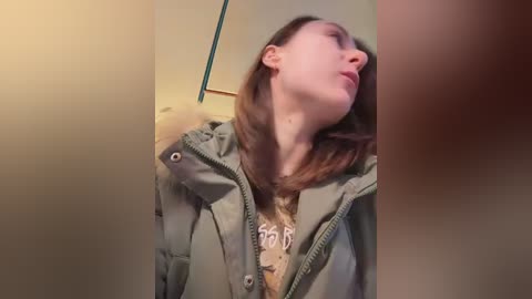 Video of a young Caucasian woman with light skin, brown hair, and fair complexion, wearing a green puffer jacket, standing in a dimly lit room with beige walls and a blue door.