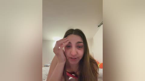 Video of a young woman with long brown hair, fair skin, and a slight smile, lying on a bed in a dimly lit room with beige walls.