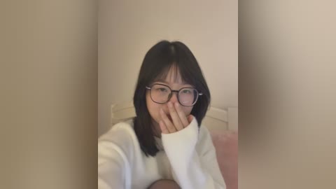 Video of an East Asian woman with short black hair, glasses, and a white sweater, covering her mouth with her hand, in a minimalist, beige-walled room.