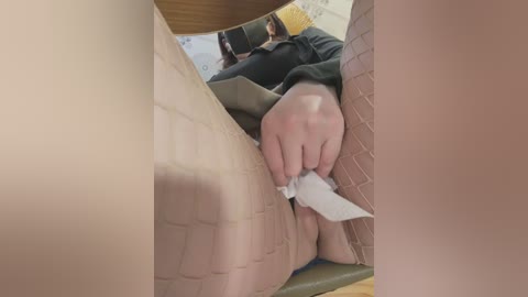 Video of a person with fair skin, wearing a yellow beanie and a black jacket, sitting on a wooden bench. Their hand is gripping a white paper, and their legs are covered in beige fishnet stockings.