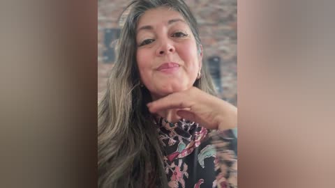 Video of a middle-aged woman with long, wavy gray hair, wearing a floral blouse, touching her chin. Brick wall background.