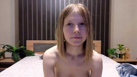 Video of a young, slender, topless, light-skinned girl with shoulder-length blonde hair, standing on a bed with striped curtains and potted plants in a modern bedroom.