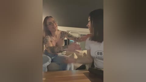 A video shows two women, one with tattoos and another with a straight bob, smiling and talking intimately in a dimly lit room with a wooden table.