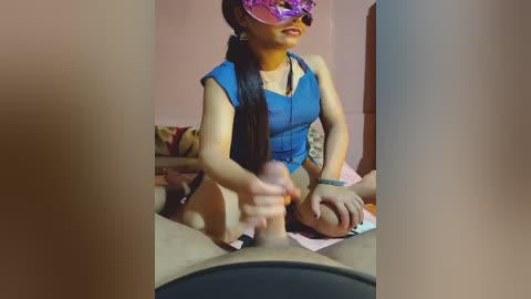 A video shows a young Asian woman with long black hair, wearing a blue sleeveless top and a pink mask, holding a large, erect penis, seated on a bed in a dimly lit room.