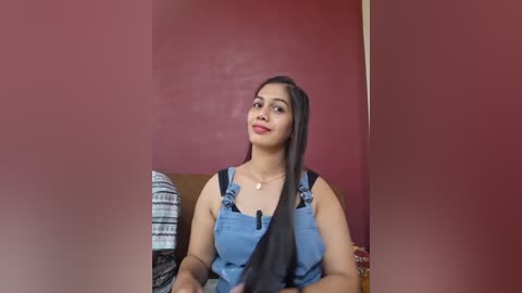 Video of a young woman with medium-brown skin and long black hair, wearing a blue denim overalls dress with black straps, seated against a red wall.