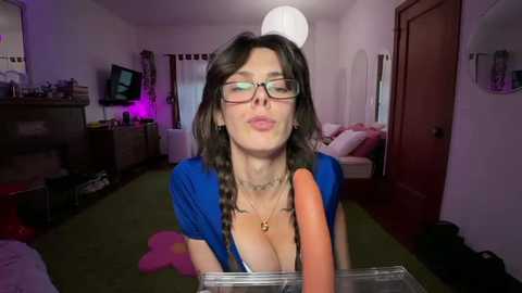 Video of a woman with glasses, long dark hair, wearing a blue top, sitting on a green carpet, holding a large carrot in a glass jar.