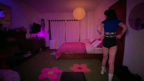 A video of a dimly lit, cozy bedroom with a person in a blue sleeveless top and black shorts, standing by the bed. Pink flower rugs adorn the floor.