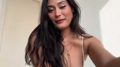 Video of a young woman with long, wavy black hair, fair skin, and a confident smile, wearing a gold necklace, against a light-colored background.