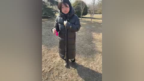 Video of a young woman with dark hair, bundled in a thick, dark winter coat, standing on a dry, grassy park path.