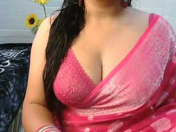 Video of a woman in a pink, see-through saree with intricate floral patterns, showcasing her medium-sized breasts in a lace bra. She has long, wet black hair and wears large, red earrings. Background includes a white wall, black patterned rug, and sunflowers.
