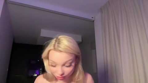 Video of a blonde Caucasian woman with fair skin and straight hair, wearing a sleeveless top, indoors with beige curtains and an air conditioner in the background.