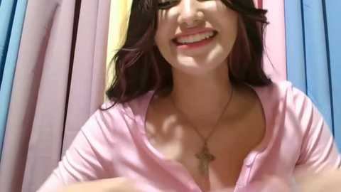 Media: Video of a smiling young woman with long, dark hair, fair skin, and medium breasts, wearing a pink, ribbed, short-sleeved top and a gold cross necklace.
