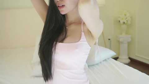 Video of an Asian woman with long black hair, fair skin, and wearing a pink camisole, stretching her arms above her head. She stands in a softly lit bedroom with white bedding, a white nightstand, and a vase of white flowers.