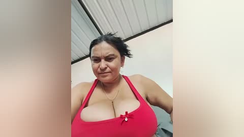 Video of a plus-size woman with dark hair and a red tank top, showcasing her large breasts. She has a neutral expression, standing in a simple room with a corrugated metal ceiling.