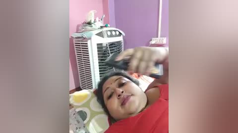Video of an Asian woman with medium skin tone and short black hair, lying on a bed with a white polka-dot pillow, wearing a red shirt. She's using a hair dryer on her hair. Background shows a white fan and purple wall.
