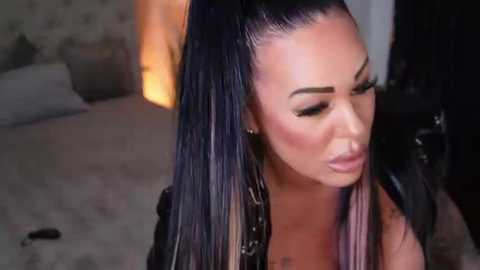 Media: A video of a woman with long, sleek black hair in a high ponytail, wearing bold makeup and a black outfit, sitting in a dimly-lit room with a cozy, warm light source in the background.