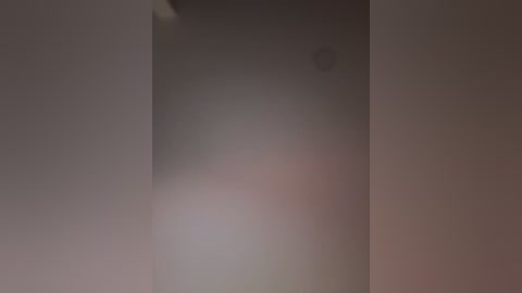 A blurred, close-up video of a dimly lit, smooth, off-white wall with a small, indistinct circular object on the upper left. The image has a soft, muted color palette, primarily shades of gray and beige, with a slight hint of pink in the center.