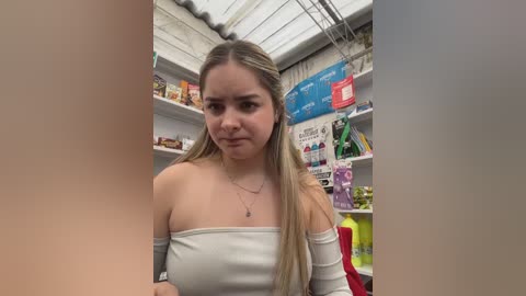 Video of a young woman with long blonde hair in a grocery store aisle, wearing a white off-shoulder top and a necklace. Shelves filled with products and signage in the background.